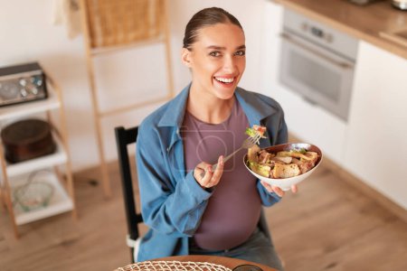 Photo for Satisfying pregnancy cravings. Happy young pregnant woman eating caesar salad, holding bowl and smiling at camera, sitting in kitchen interior - Royalty Free Image