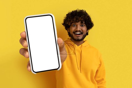 Foto de Bonita oferta online. fresco alegre casual guapo joven indio mostrando gran teléfono inteligente con pantalla blanca en blanco en la mano, aislado en el fondo del estudio amarillo, maqueta, espacio de copia - Imagen libre de derechos