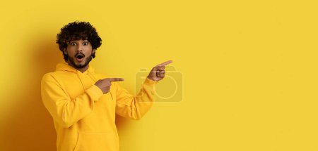 Photo for Amazed shocked handsome millennial eastern guy wearing yellow hoodie pointing at copy space for advertisement or text, showing great deal or offer, isolated on yellow background, mockup, web-banner - Royalty Free Image