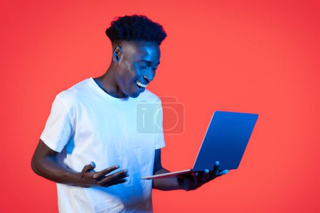 Photo for Online assistant, chatbot, artificial intelligence and daily life. Happy amazed young black man wearing white t-shirt using modern computer pc laptop and gesturing over red background in neon light - Royalty Free Image