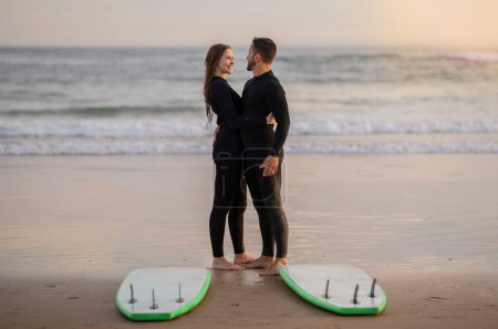 Foto de Pareja de surfistas románticos teniendo una cita en la playa, abrazándose y sonriéndose mutuamente, feliz hombre y mujer del milenio vistiendo trajes de neopreno cerca de sus tablas de surf, abrazándose y divirtiéndose - Imagen libre de derechos