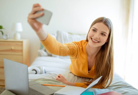 Foto de E-Learning Fun. Joven estudiante Lady aprendizaje en el ordenador portátil y hacer selfie celebración Smartphone posando en la cama en el acogedor dormitorio en casa. Redes sociales y gadgets en el concepto de la edad adolescente - Imagen libre de derechos