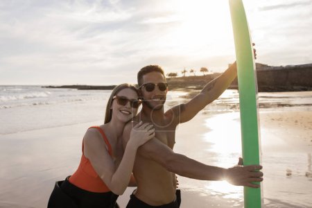 Photo for Summer Adventures. Cheerful Young Surfers Couple Posing On The Beach At Sunset, Happy Millennial Man And Woman In Stylish Sunglasses Surfing Together And Having Fun, Looking At Camera - Royalty Free Image