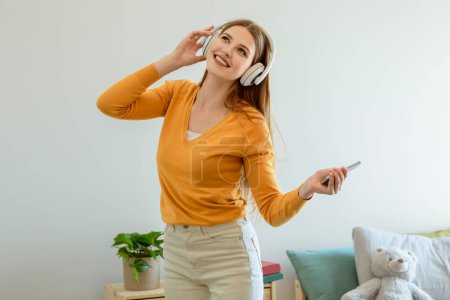 Photo for Musical Fun. Young Cheerful Blonde Lady With Smartphone Wearing Earphones, Listening To Music Online And Dancing At Home Interior, Standing In Modern Bedroom, Relaxing On Weekend - Royalty Free Image