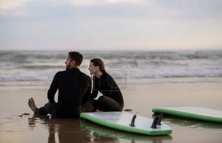 Photo for Young Couple In Wetsuits Relaxing On The Beach After Surfing, Happy Romantic Man And Woman Sitting On Sand Near Their Surfboards, Millennial Lovers Chilling On Seashore Together, Copy Space - Royalty Free Image