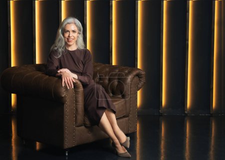 Photo for Successful Business Lady. Smiling Mature Businesswoman In Formal Dress Sitting In Brown Leather Armchair, Posing Looking At Camera At Dark Luxury Interior. Career Success And Entrepreneurship - Royalty Free Image