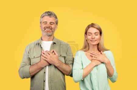 Photo for Positive european middle aged couple with closed eyes making gesture of gratitude, putting hands to chest, posing on yellow studio background. People emotions, thank, love sign, ad and offer - Royalty Free Image