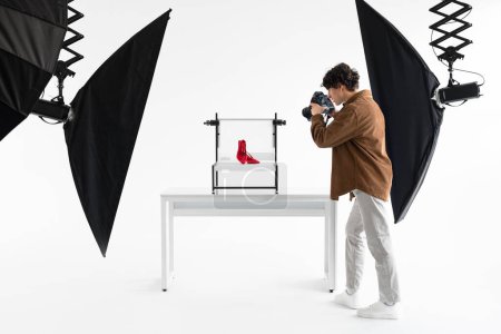 Photo for Professional male content maker making photoshoot for shop, taking photos of elegant red shoes, working in modern photostudio with lighting equipment, full length shot - Royalty Free Image