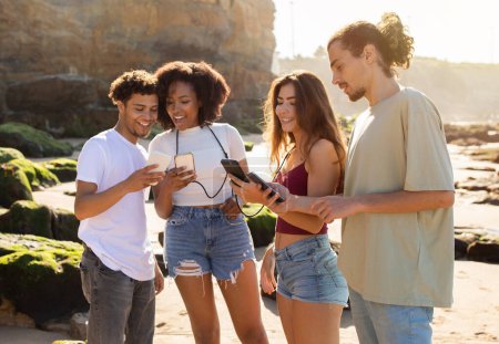 Photo for Cheerful millennial international people students enjoy summer holidays, have fun, chatting on smartphones on beach. Blog, social network app, active lifestyle, free time at weekend together - Royalty Free Image