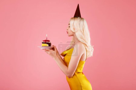 Foto de Mujer joven y feliz vestido y sombrero de fiesta soplando vela en el pedazo de pastel, celebrando su cumpleaños, de pie aislado en el fondo del estudio rosa, vista lateral - Imagen libre de derechos