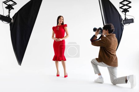 Téléchargez les photos : Photoshoot de mode. Photographe professionnel prenant des photos de beau modèle féminin en robe rouge élégante, posant sur fond blanc dans un studio photo avec un équipement d'éclairage moderne - en image libre de droit