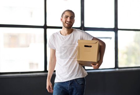 Téléchargez les photos : L'emploi. Happy Middle Eastern Employee Guy Holding Cardborad Box Avec Belongings Obtenir un nouvel emploi debout dans occasionnel dans l'intérieur de bureau moderne, souriant à la caméra. Croissance de carrière, réinstallation - en image libre de droit