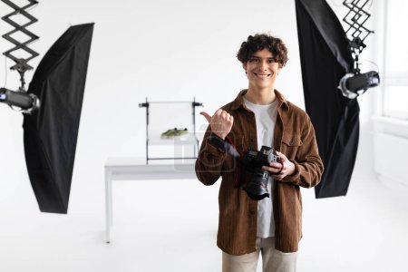 Téléchargez les photos : Portrait de photographe masculin heureux en tenue décontractée tenant l'appareil photo numérique et pointant du doigt tout en faisant des séances photo de contenu, appréciant son travail au studio de photo - en image libre de droit
