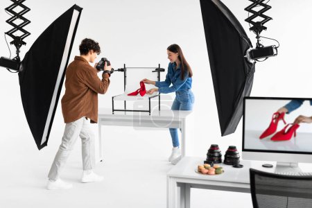 Téléchargez les photos : Équipe professionnelle de photographe homme et assistante féminine prenant des photos de chaussures rouges élégantes, travaillant dans un photostudio moderne, faisant photoshoot de contenu - en image libre de droit