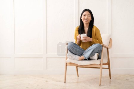 Foto de Alegre bonita de pelo largo millennial asiático mujer usando casual traje sentado en sillón, sosteniendo blanco taza, bebiendo café y sonriendo en cámara, hogar interior, copia espacio - Imagen libre de derechos