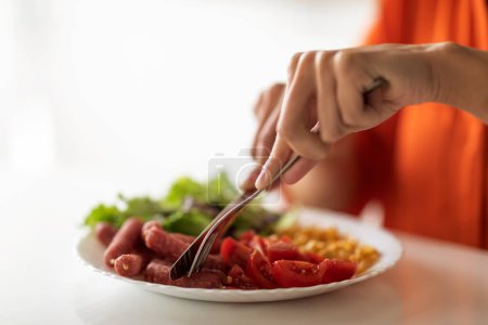 Foto de Mujer negra irreconocible comiendo sabroso almuerzo mientras está sentada en la mesa en la cocina, joven mujer afroamericana usando tenedor y cuchillo, disfrutando de la comida, teniendo ensalada fresca, salchichas y tomates, recortados - Imagen libre de derechos