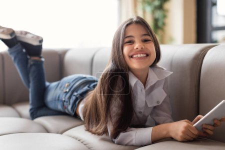 Photo for Mobile Offer. Cheerful Preteen Kid Girl Using Phone And Smiling To Camera Posing On Sofa At Home. Child Scrolling Social Media, Engaging In Online Entertainment On Cellphone. Children And Gadgets - Royalty Free Image
