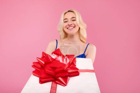 Photo for Great idea for present. Beautiful charming young woman in dress giving you big wrapped gift with red bow and smiling, posing over pink studio background - Royalty Free Image