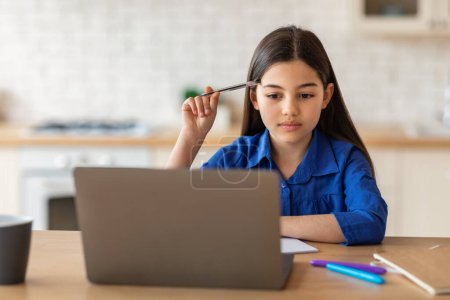Téléchargez les photos : E-Learning. Pensive petite écolière étudiant avec ordinateur portable à la maison, tenant le stylo et regardant l'ordinateur faire des devoirs. Kid Schooler penser à un projet éducatif en ligne - en image libre de droit