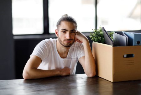 Photo for Dismissal. Fired Arabic Employee Man Sitting At Table With His Belongings In Cardboard Box At Workplace, Looking At Camera Posing In Modern Office Interior. Unemployment Problem Concept - Royalty Free Image