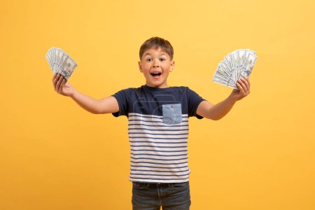 Photo for Thrilled child cute handsome caucasian boy wearing casual outfit holding money cash dollar banknotes in both hands and exclaiming, isolated on yellow background. Pocket money for kids - Royalty Free Image
