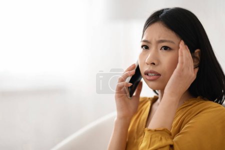 Photo for Closeup of anxious worried beautiful millennial asian woman in comfy casual outwear sitting on couch, having phone conversation, touching head, looking at copy space for ad, have troubles - Royalty Free Image