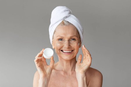 Photo for Happy senior woman with towel on head holding jar with moisturising anti-aging cream, pampering herself and smiling at camera, posing on grey background - Royalty Free Image