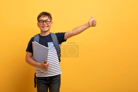 Téléchargez les photos : Joyeux garçon écolier caucasien joyeux avec un sac à dos derrière le dos et des livres à la main montrant le pouce levé et souriant, isolé sur fond jaune, espace de copie. Grande offre pour les écoliers - en image libre de droit