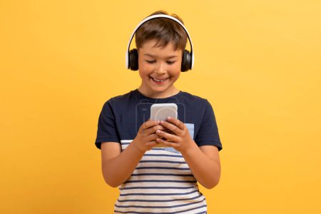 Photo for Cheerful cute handsome school aged boy in casual outfit using cell phone and wireless headphones, isolated on yellow background. Happy kid watching video content, playing games online - Royalty Free Image