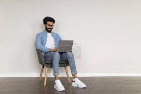 Téléchargez les photos : Homme indépendant indien réussi utilisant un ordinateur portable, travaillant en ligne tout en étant assis dans un fauteuil à l'intérieur, Homme millénaire oriental naviguant sur Internet sur ordinateur ou écrire l'article pour le blog, Copier l'espace - en image libre de droit
