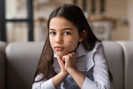 Photo for Unhappy Preteen Girl Looking At Camera With Sad Expression Leaning Head On Hands, Posing At Modern Home Interior. Portrait Of Depressed Middle Eastern Child Feeling Depressed And Lonely - Royalty Free Image