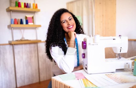 Téléchargez les photos : Conception de vêtements. Femme couturière latine professionnelle assise près de la machine à coudre posant sur le lieu de travail dans un atelier de tailleur moderne à l'intérieur, souriant à la caméra. Tourné de Dressmaker réussie au travail - en image libre de droit