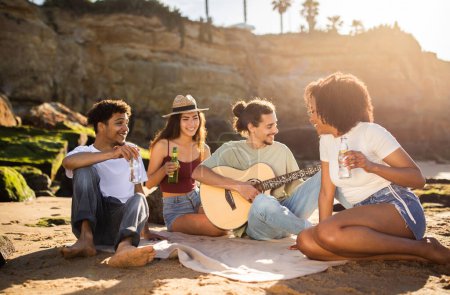Foto de Sonriendo millennial multiétnicos estudiantes con botellas, tocando la guitarra, disfrutar de vacaciones de verano, picnic, divertirse en la playa. Música estilo de vida, tiempo libre, fiesta de fin de semana juntos - Imagen libre de derechos