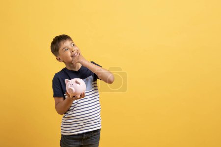 Photo for Kids spendings, pocket money. Happy excited handsome school aged child caucasian boy in casual outfit holding piggy bank, touching his face and looking at copy space, isolated on yellow background - Royalty Free Image