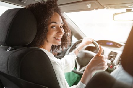 Téléchargez les photos : Jeune femme du Moyen-Orient souriante conduisant une nouvelle voiture, montrant le pouce levé geste assis à l'intérieur de son véhicule de luxe, vue depuis le siège arrière. Portrait de dame heureuse propriétaire d'automobile - en image libre de droit