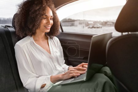 Téléchargez les photos : Transfert confortable. Femme d'affaires souriante du Moyen-Orient utilisant un ordinateur portable sur le siège arrière dans une voiture moderne, travaillant en ligne avec un ordinateur tout en voyageant dans un véhicule, allant à une réunion d'affaires. Vue latérale - en image libre de droit
