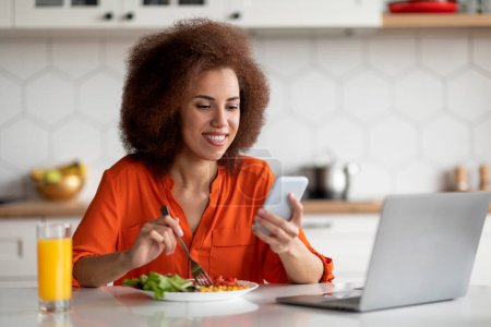 Photo for Smiling black woman using smartphone and laptop while having lunch in kitchen at home, cheerful african american female sitting at table, browsing new app on mobile phone and eating food, free space - Royalty Free Image