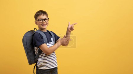 Foto de Feliz niño caucásico alegre colegial con mochila detrás de la espalda apuntando al espacio de copia para la publicidad y sonriente, aislado sobre fondo amarillo. Gran oferta para los escolares - Imagen libre de derechos