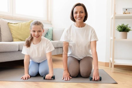 Photo for Positive millennial caucasian woman in sportswear and teenage daughter training with dumbbells together in living room interior, enjoy sports at home. Body care and fit, active lifestyle - Royalty Free Image