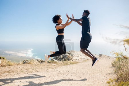 Photo for Young black couple jumping and giving high five, enjoying workout together in morning on rocks at ocean, outdoors. Sports at summer, teamwork and good result - Royalty Free Image
