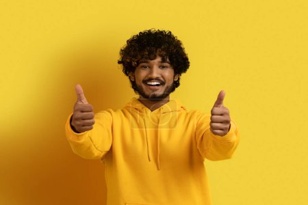 Photo for Cheerful handsome eastern guy wearing yellow hoodie showing thumb ups and smiling at camera, happy customer like or recommend something nice, isolated on yellow background - Royalty Free Image