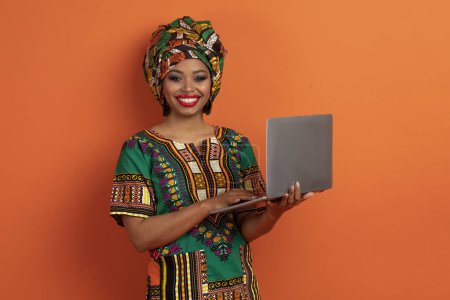 Photo for Chatbot, online assistant, artificial intelligence. Happy positive young african lady in national clothing digital nomad using modern pc laptop on orange studio background - Royalty Free Image