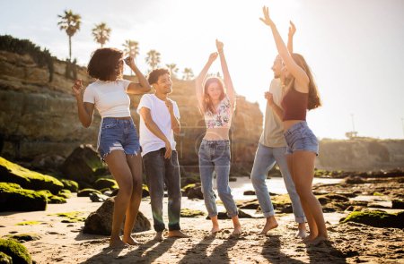 Foto de Alegre millennial negros, árabes, caucásicos estudiantes disfrutan de vacaciones de verano, divertirse, bailar, levantar la mano en la playa. Fiesta junto con amigos, viaje, estilo de vida activo, tiempo libre en fin de semana - Imagen libre de derechos