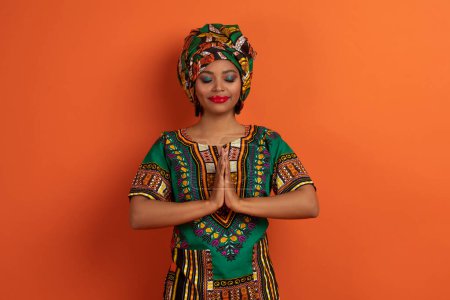 Foto de Tranquila calma atractiva joven mujer negra vistiendo traje africano tradicional cogido de la mano en gesto namaste sobre el pecho, meditando con los ojos cerrados o rezando sobre fondo naranja - Imagen libre de derechos