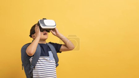 Foto de Colegial en traje casual con mochila a sus espaldas usando gafas VR aisladas sobre fondo amarillo, experiencia de realidad virtual, espacio para copiar, banner. Tecnologías modernas en la educación infantil - Imagen libre de derechos