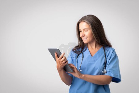 Téléchargez les photos : Heureux médecin femme européenne senior en uniforme bleu tapant sur tablette numérique, posant isolé sur fond gris clair, espace libre. App pour le traitement des maladies, le diagnostic, les soins de santé - en image libre de droit