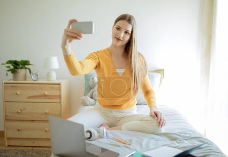 Foto de Diversión en redes sociales. Mujer joven haciendo selfie en el teléfono inteligente, mientras que el aprendizaje en línea y hacer la tarea en el ordenador portátil en el acogedor dormitorio interior. Concepto de Estudiancia Moderna - Imagen libre de derechos