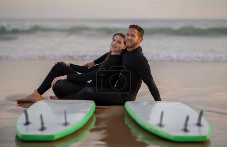 Photo for Romantic Young Couple Wearing Wetsuits Posing With Surfboards On The Beach, Happy Millennial Man And Woman Surfers Sitting On Seashore And Looking At Camera, Enjoying Surfing Together - Royalty Free Image