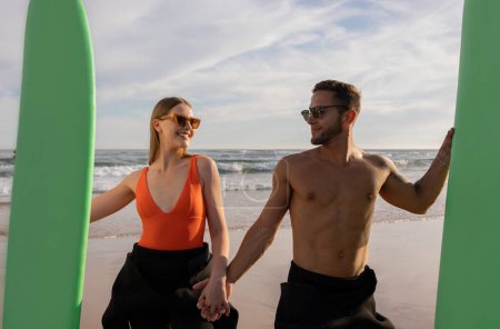 Foto de Retrato de hermosa pareja de jóvenes surfistas con tablas de surf posando en la playa, amantes felices con gafas de sol con estilo sosteniendo las manos y mirándose unos a otros, divirtiéndose mientras navegan juntos - Imagen libre de derechos