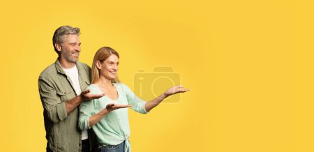 Téléchargez les photos : Époux d'âge moyen joyeux pointant de côté l'espace libre, homme et femme heureux manifestant leur place pour le texte ou le design, debout sur fond de studio jaune, panorama - en image libre de droit